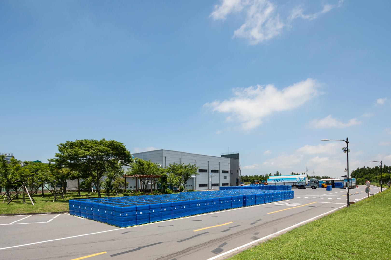 Blue pallets of bottled water stacked in rows outside the Samdasoo plant, with a Samdasoo-logo truck waiting in the foreground